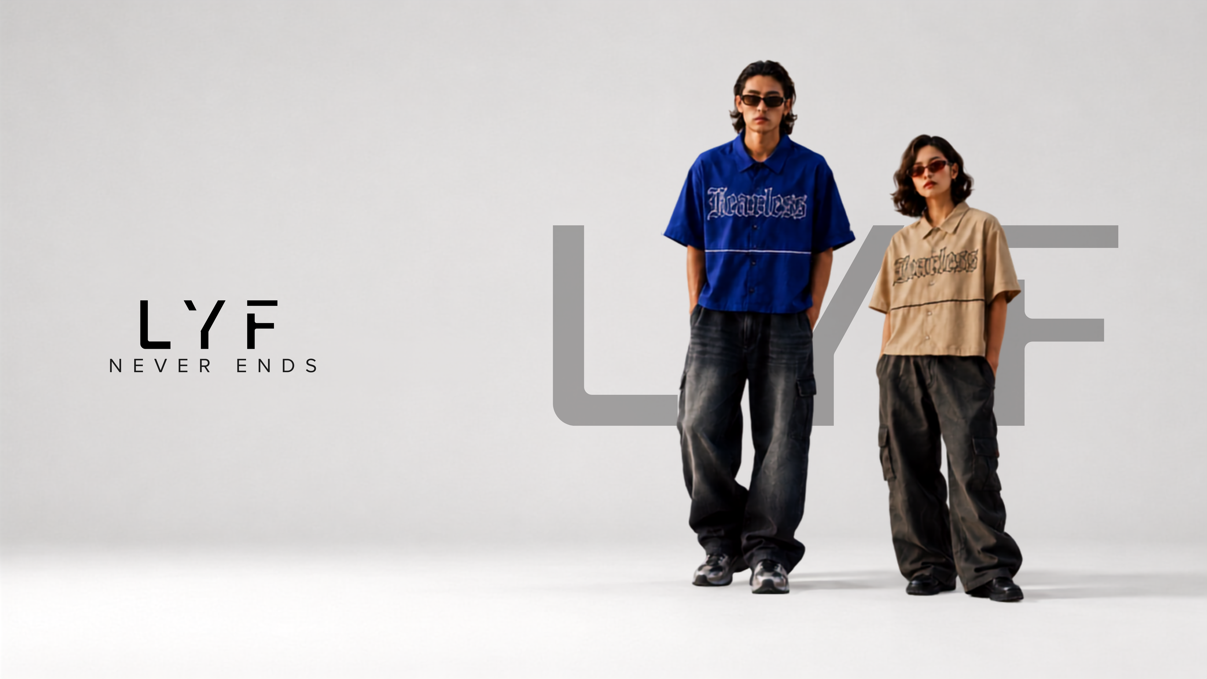 Two models standing in a studio wearing oversized LYF Never Ends graphic t-shirts and baggy dark pants. The model on the left wears a blue t-shirt and the model on the right wears a beige t-shirt.